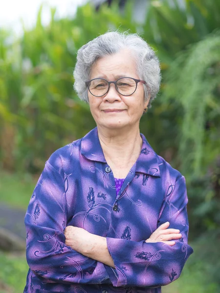 Portrait of a senior woman with short gray hair smiling, arms crossed and looking at the camera standing in a garden. Concept of aged people and relaxation.