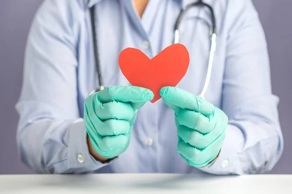 Hand of a doctor showing a red paper heart shape while sitting on a chair with gray background in the hospital or clinic. Close-up photo. Space for text. Medical and encouragement concept.