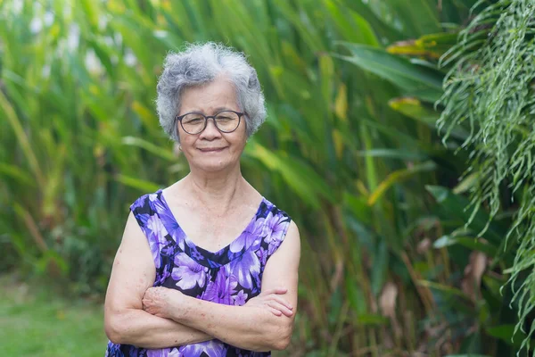 Portrait of a senior woman with short gray hair, wearing glasses, smiling and looking at the camera while standing in a garden. Aged people and relaxation concept.