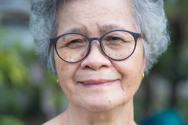 Portrait of an elderly woman with short gray hair, wearing glasses, smiling, and looking at the camera while standing in a garden. Aged people and relaxation concept.