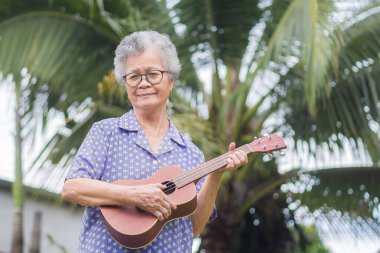 Hindistan cevizi ağacının arka planında bahçede dikilirken ukulele çalan kısa gri saçlı mutlu son sınıf kadını. Emekli olduktan sonra hayatın tadını çıkar. Yaşlılar ve rahatlama kavramı.
