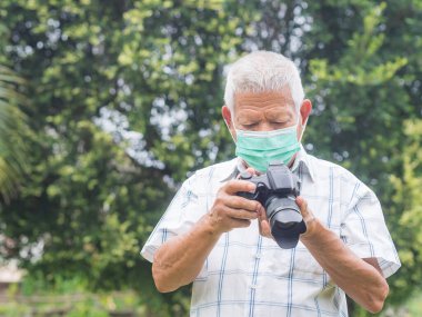 Yüz maskesi takan ve dijital kamera kullanan son sınıf bir adamın açık hava portresi. Profesyonel fotoğrafçı. Emekli olduktan sonra Asyalı yaşlı adamın mutluluğu. Yaşlılar ve fotoğrafçılık konsepti.