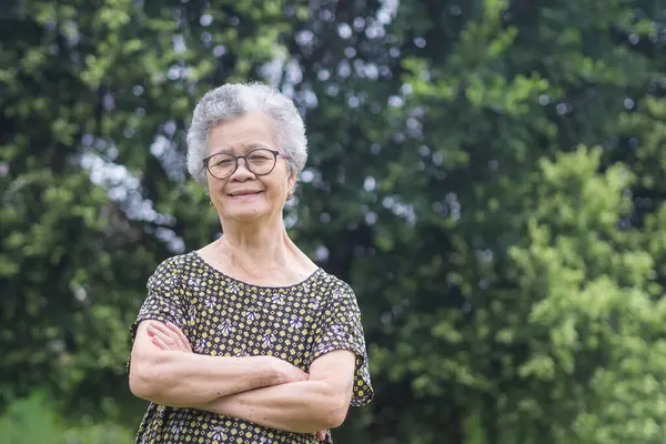 Portrait of a senior woman with short gray hair, wearing glasses, smiling, arms crossed, and looking at the camera while standing in a garden. Aged people and relaxation concept.