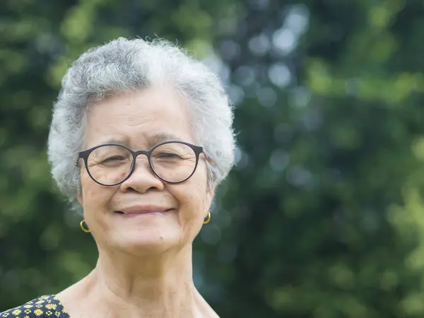 Portrait of a senior woman with short gray hair, wearing glasses, smiling, and looking at the camera while standing in a garden. Aged people and relaxation concept.