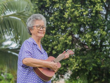 Bahçede dikilirken ukulele çalan kısa gri saçlı yaşlı bir Asyalı kadının portresi. Emekli olduktan sonra hayatın tadını çıkar. Yaşlılar ve rahatlama kavramı.