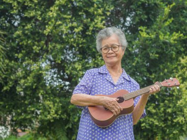 Kısa gri saçlı, ukulele çalan, gülümseyen ve bahçede dikilirken kameraya bakan mutlu yaşlı kadın. Emekli olduktan sonra hayatın tadını çıkar. Yaşlılar ve rahatlama kavramı.