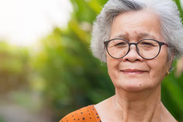Portrait of a senior woman with short gray hair, wearing glasses, smiling, and looking at the camera while standing in a garden. Aged people and relaxation concept.