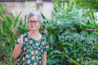 Kısa gri saçlı mutlu bir Asyalı kadın, gözlük takıyor, başparmağını kaldırıyor ve bahçede dikilirken kameraya bakıyor. Yaşlılar ve rahatlama konsepti.