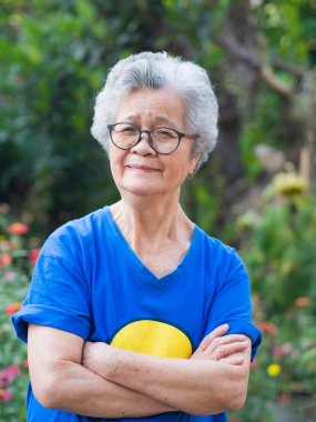 Mutlu, kısa gri saçlı, gülümseyen, kollarını kavuşturup bahçede duran kameraya bakan son sınıf öğrencisi bir kadın. Yaşlılar ve rahatlama kavramı.