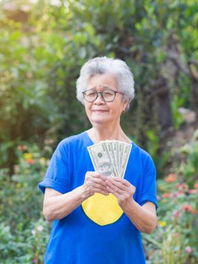Kısa gri saçlı neşeli yaşlı kadın Amerikan dolarlarını elinde tutuyor ve bahçede dururken gülümseyerek kameraya bakıyor..