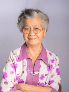 Kısa gri saçlı, güzel Asyalı kadın gözlük takıyor ve gri arka planda dururken kameraya gülümsüyor. Yaşlılar ve sağlık hizmetleri kavramı.