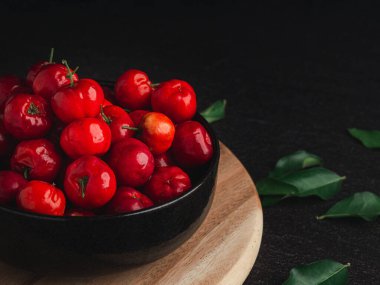 Olgun kırmızı acerola kirazları, tahtadan kesme tahtasındaki seramik bir kasede, koyu yeşil yapraklı bir masa üzerinde meyve verir. Yüksek C vitamini ve antioksidan meyveler. Yakın plan fotoğraf. Sağlıklı meyve kavramı.