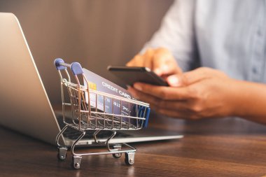 Ofiste otururken arka planı bulanıklaştıran akıllı telefon kullanan bir adamla ahşap bir masada mini bir alışveriş arabası. İş ve e-ticaret kavramı