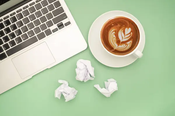 Working desk table concept. Top view of a laptop, crumpled paper, and a white coffee cup isolated on a green background.