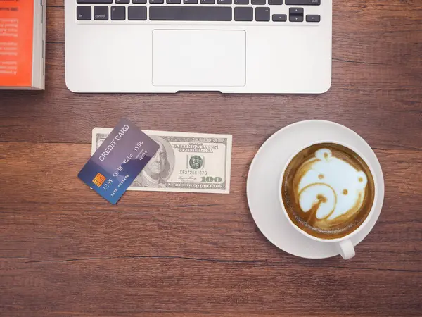 Working desk table concept. Top view of a mockup blue credit card over on dollar banknotes, a book, a laptop, and a coffee cup on a wooden table in the office. Flat lay, space for text.