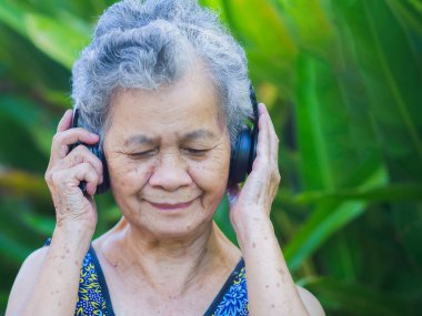 Kısa gri saçlı, kablosuz kulaklık takan neşeli son sınıf öğrencisi bir kadın bahçede dururken gülümseyerek en sevdiği şarkıyı dinliyor. Yaşlılar ve rahatlama konsepti.