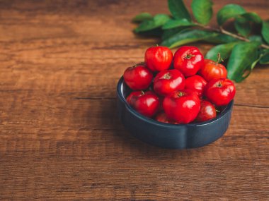 Olgun kırmızı acerola, seramik bir kasede meyve ve ahşap bir masada yeşil yapraklar. Yüksek C vitamini ve antioksidan meyveler. Yakın plan fotoğraf. Mesaj için yer var. Sağlıklı meyve kavramı.