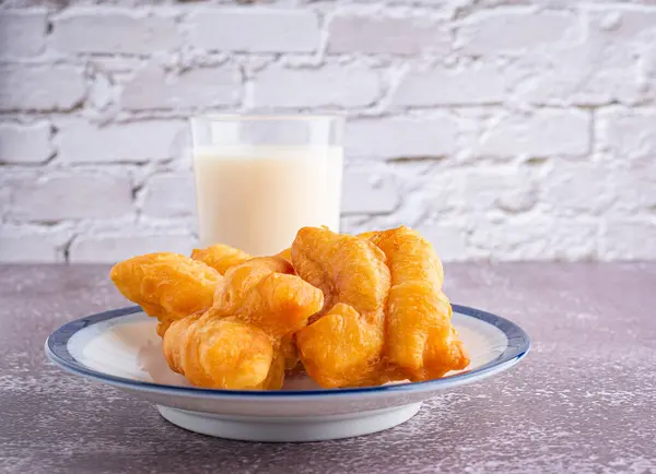 Close-up of deep-fried dough sticks or Chinese doughnut sticks on white plate and a glass of soybean milk on gray stone background with white brick wall. Space for text.