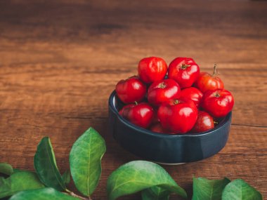 Kase içinde kırmızı acerola kirazı ahşap masa arkasında yeşil yapraklar. Yüksek C vitamini ve antioksidan meyveler. Üst Manzara. Sağlıklı meyve kavramı.