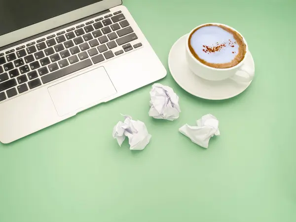Working desk table concept. Top view of a laptop, crumpled paper, and a white coffee cup isolated on a green background.