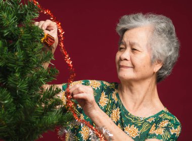 Stüdyoda kırmızı bir arka planda otururken Noel ağacını süsleyen neşeli son sınıf kadını. Mesaj için yer var. Seçici odaklanma. Yaşlılar ve Noel kavramı ve mutlu yeni yıl festivali.