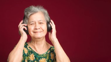 Kırmızı arka planda dururken gülümseyerek en sevdiği şarkıyı dinlemek için kısa gri saçlı, kablosuz kulaklık takan neşeli, yaşlı Asyalı kadın. Yaşlılar ve rahatlama konsepti.