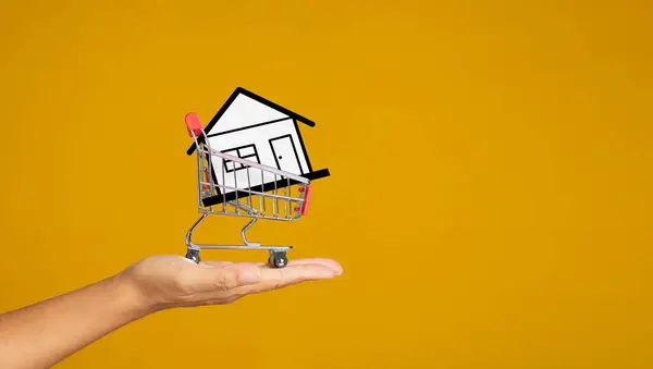 Home loan and real estate investment concept. Close-up of a mockup white paper home in a mini shopping trolley on the palm with a yellow background. Property insurance and security