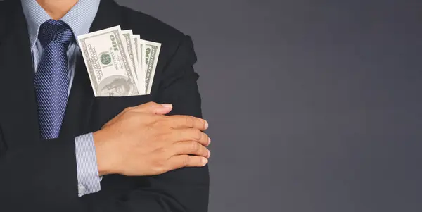 Business, finance, payday loan concept. Businessman wearing a suit with US dollar banknotes in a suit pocket and arms crossed while standing with a gray background with space for text.
