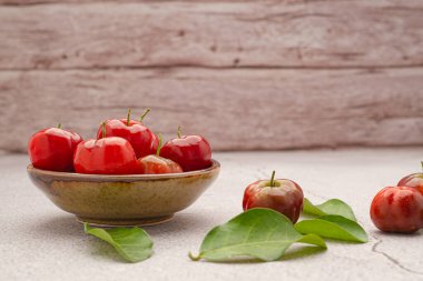 Sağlıklı meyve kavramı. Olgun kırmızı acerola, seramik bir kasede meyveler ve ahşap duvarlı çimento zeminde yeşil yapraklar. Yüksek C vitamini ve antioksidan meyveler. Metin için boşluk.