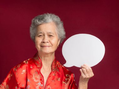 Konuşma balonu konsepti. Geleneksel bir Cheongsam Qipao elbisesi giymiş, elinde boş bir kağıt baloncukla stüdyoda otururken kameraya gülümseyen yaşlı bir Asyalı kadının portresi.