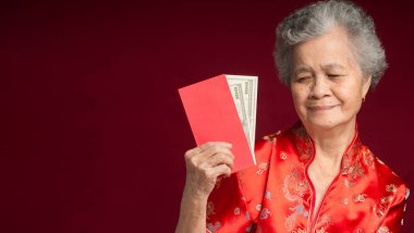 Çin Yeni Yıl konsepti. Geleneksel Cheongsam Qipao elbisesi giymiş yaşlı bir Asyalı kadının portresi. Angpao 'yu elinde ABD Doları banknotuyla stüdyoda kırmızı arka planda dururken tutuyor.