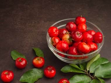 Cam bir kasede olgun kırmızı acerola kirazları ve masaya saçılmış yeşil yapraklar. Yüksek C vitamini ve antioksidan meyveler. Mesaj için yer var. Yakın plan. Sağlıklı meyve kavramı