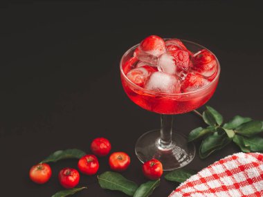 Siyah arka planda kiraz suyu olan şarap bardağı. Soğuk içecek ferahlatıcı. Yaz partisi. Masadaki olgun kırmızı acerola kirazı ve yeşil yapraklar. Yüksek C vitamini meyveleri