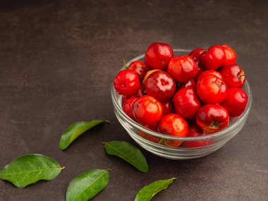 Cam bir kasede olgun kırmızı acerola kirazları ve masaya saçılmış yeşil yapraklar. Yüksek C vitamini ve antioksidan meyveler. Mesaj için yer var. Yakın plan. Sağlıklı meyve kavramı