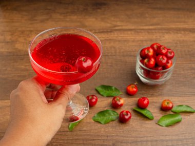 Kiraz suyuyla birlikte bir şarap bardağını tutan el. Cam kirazlar ve ahşap bir masada yeşil yapraklar içinde olgun kırmızı acerola. Yüksek C vitamini ve antioksidan meyve. Yakın plan fotoğraf.