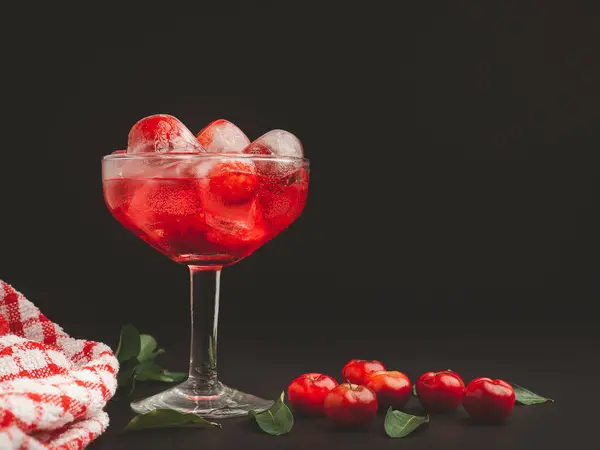 stock image Wine glass with cherry juice on a black background. Refreshing cold drink. Summer party. Ripe red acerola cherries and green leaves on a table. High vitamin C fruits