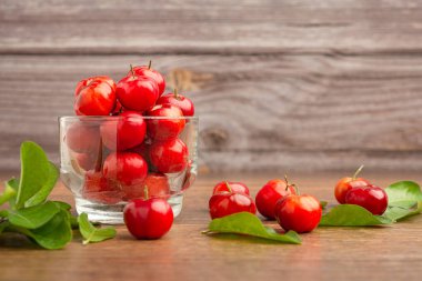 Cam kaplı olgun kırmızı acerola kirazları ve ahşap duvarlı bir masa üzerine serpilmiş yeşil yapraklar. Yüksek C vitamini ve antioksidan meyveler. Mesaj için yer var. Yakın plan. Sağlıklı meyve kavramı