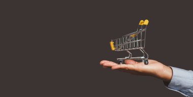 İş ve e-ticaret kavramı. Stüdyoda gri arka planda dikilirken palmiye insanlarının üzerinde boş sarı bir alışveriş arabasının yakın çekimi. Yan görüş. Metin için boşluk.