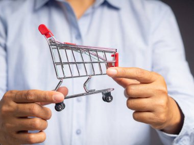 İş ve e-ticaret kavramı. Gri bir arka planda dururken küçük bir alışveriş arabasını tutan ellerin yakın çekimi.