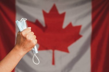 COVID-19 ya da Coronavirus 'un sonu. Kanada bayrağının arka planında bir yumrukta tıbbi maskenin yakın çekimi. Normal hayata dönmek karantinanın sonu.
