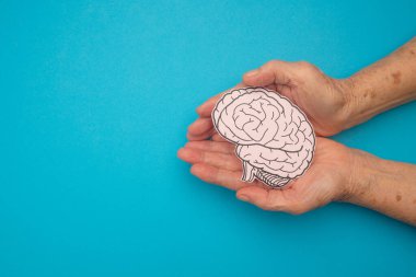 Mavi arka planda, avuç içi yaşlı bir kadının kağıdından yapılmış bir beyin şekli. Alzheimer, Parkinson hastalığı, bunama, felç, nöbet ya da akıl sağlığı. Nöroloji ve Psikoloji