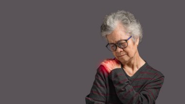 Yaşlı kadın gri arka planda dururken omuz ağrısı çekiyor. Yaşlılar ve sağlık konsepti