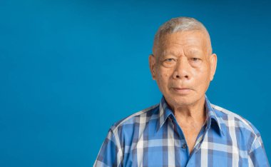 Stüdyoda mavi arka planda dikilirken kameraya gülümseyen mavi gömlekli yaşlı Asyalı bir adamın portresi. Mesaj için yer var. Yaşlılar ve sağlık konsepti
