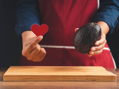 Hand holding a red heart shape symbol with fresh avocado while standing in the kitchen. Healthy fruit and food eating concept. Close-up photo clipart