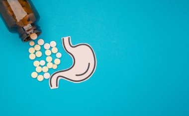 Kağıttan yapılmış mide şekli ve mavi arka planda şişesi olan haplar. Mide ağrısına regl dönemi, mide ülseri, apandisit ya da sindirim sistemi hastalığı neden olur.