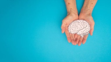 Mavi arka planda, avuç içi yaşlı bir kadının kağıdından yapılmış bir beyin şekli. Alzheimer, Parkinson hastalığı, bunama, felç, nöbet ya da akıl sağlığı. Nöroloji ve Psikoloji