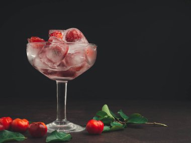 Buzlu taze kirazlı şarap bardağı. Soğuk içecek ferahlatıcı. Yaz partisi. Masadaki olgun kırmızı acerola kirazı ve yeşil yapraklar. Yüksek C vitamini meyveleri