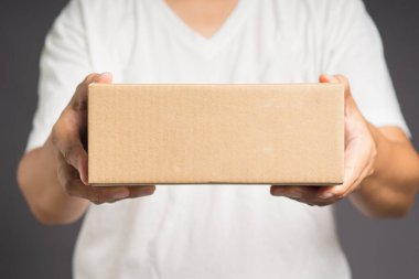 Gri arka planda duran beyaz giysili bir adam karton bir kutu tutuyor.