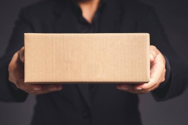 Siyah takım elbiseli bir işadamı gri arka planda dururken bir karton kutuyu işaret ediyor. Mesaj için yer var. Yakın plan fotoğraf. İş, lojistik ve e-ticaret kavramı