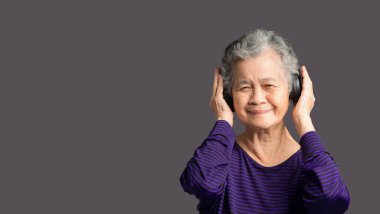 Neşeli, yaşlı Asyalı, kablosuz kulaklık takan, gülümseyen ve gri arka planda dururken kameraya bakan favori bir şarkıyı dinleyen bir kadın. Yaşlılar ve rahatlama konsepti
